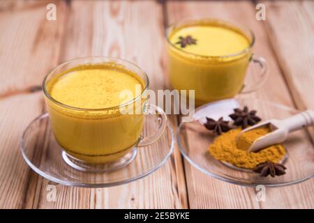 Becher mit goldenem Milchgetränk und Kurkuma-Pulver mit Holzlöffel auf Holzhintergrund. Stockfoto