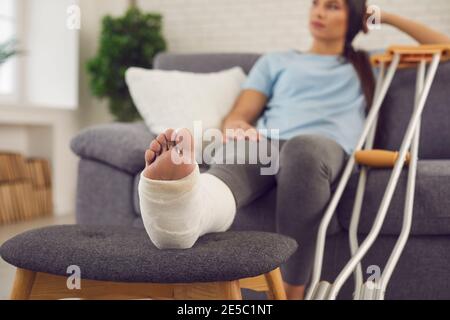 Junge Frau mit verletzten Bein sitzt auf der Couch zu Hause Mit zwei Krücken daneben Stockfoto