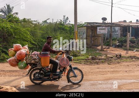 Nandakeshwar, Karnataka, Indien - 7. November 2013: Ambulanter Händler auf dem Motorrad verkauft bunte Bälle für Kinder Fahrten auf Feldweg mit grünen Foli Stockfoto