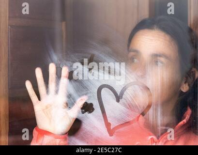 Frau mittleren Alters, die aus dem Fenster schaut. Kondensation am Fenster. Stockfoto