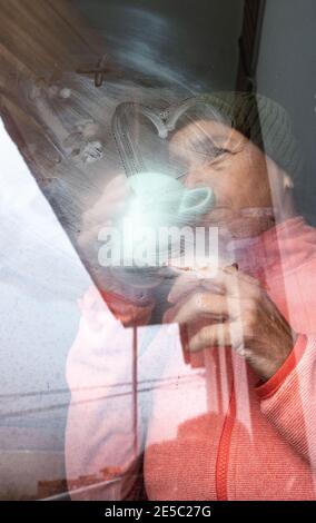 Frau mittleren Alters mit einer Tasse Kaffee, die an einem kalten Tag aus dem Fenster schaute. Stockfoto