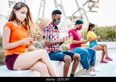 Multikulturelle Freunde mit OP-Maske mit Tracking-App auf Handy Smartphones - gelangweilte junge Menschen am Riesenrad Stockfoto