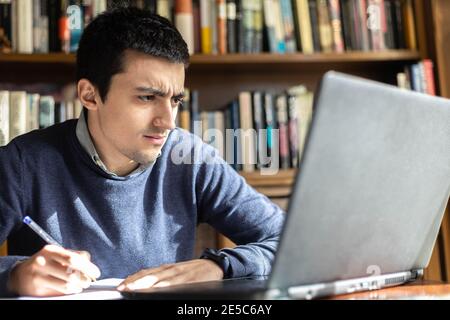 Selektiver Fokus eines jungen Mannes überrascht von den Noten Er hat auf dem Computer während der Arbeit oder Online-Studium Und Notizen in einem Notizbuch auf Stockfoto