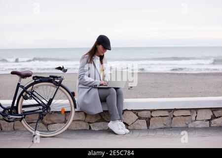 Foto einer freiberuflichen Mitarbeiterin, die im Freien auf einem Laptop arbeitet, der mit dem öffentlichen WLAN verbunden ist. Online arbeitet, während sie Zeit am Meer verbringt Stockfoto