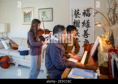 Ein Vater und ein Onkel sitzen am Klavier, während das Mädchen spielt Geige hinter ihnen Stockfoto