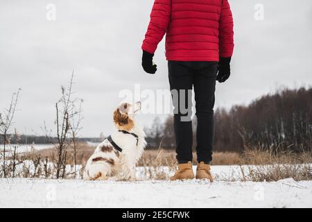 Selbstbewusster glücklicher Hund mit geschlossenen Augen sitzt neben seinem Besitzer im Freien an einem gefrorenen verschneiten Fluss. Wandern, Wandern mit Haustieren im Winter - Mann im roten Winter Stockfoto