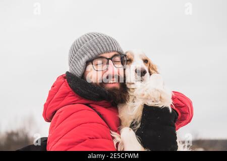 Ein glücklicher Rüde umarmt seinen Spaniel-Hund bei einem Spaziergang. Verbringen Sie Zeit mit Haustieren im Freien, Wandern im Winter, Freundschaft und Zuneigung mit Hunden Stockfoto