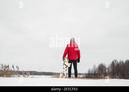 Ein Hund steht neben seinem Besitzer draußen an einem gefrorenen verschneiten Fluss. Wandern, Wandern mit Haustieren im Winter - Mann in roter Winterjacke genießt Freizeit mit Stockfoto