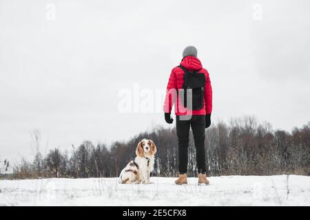 Ein Hund sitzt neben seinem Besitzer im Freien. Wandern, Wandern mit Haustieren im Winter - Mann in roter Winterjacke genießt Freizeit mit seinem Hund Stockfoto