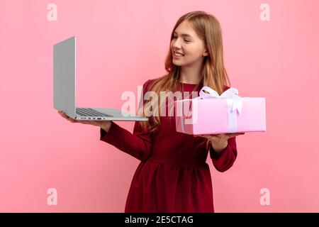 Happy attraktive junge Frau in eleganten roten Kleid mit Geschenkbox nutzt Laptop-Computer, auf rosa Hintergrund, Konzept des Valentinstag, Einkaufen Stockfoto