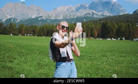 Mittellange Aufnahme einer Frau, die einen Videoanruf aus einem wunderschönen Naturpark vor den Bergen macht. Sie teilt Eindrücke von ihrer Reise. Stockfoto