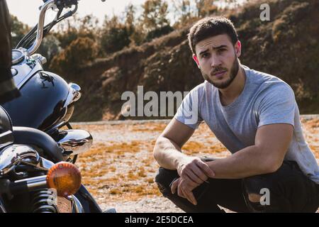 Der junge Biker hockend neben seinem Motorrad und schaut auf die Kamera Stockfoto