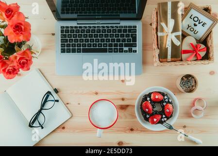 Arbeitsplatz von oben mit Laptop, Notebook, Brille und süßem Valentinstag-Überraschungsfrühstück. Tasse Latte Kaffee, Bagel mit Erdbeeren und Stockfoto
