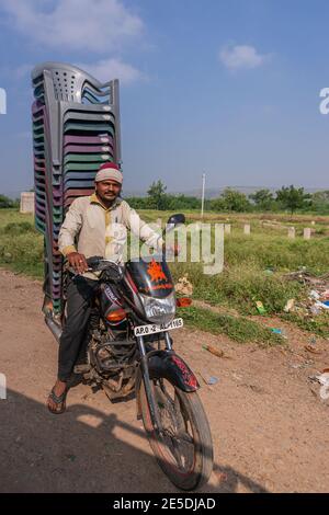 Pattadakai, Karnataka, Indien - 7. November 2013: Junger Mann auf dem Motorrad transportiert einen Haufen Plastikstühle. Grüne landwirtschaftliche Flächen im Rücken unter BL Stockfoto