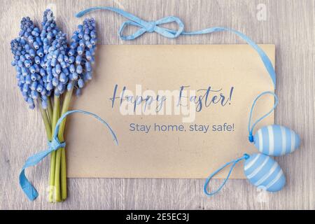 Text Frohe Ostern, zu Hause bleiben, sicher bleiben auf Handwerk Papierkarte eingerahmt von blauen Bändern, Ostereiern und Traubenhyazinthe Blumen Stockfoto