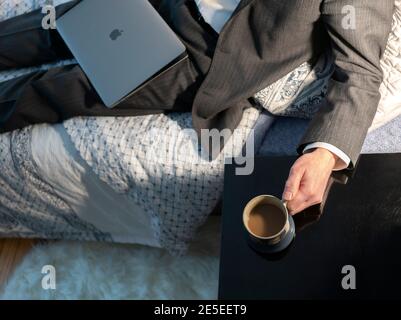 Arbeit von zu Hause Konzept Mann trägt einen Anzug auf dem Bett mit Apple Laptop Computer Hand hält Tasse Kaffee. Siehe 2E5EET7 No Brand MR Stockfoto