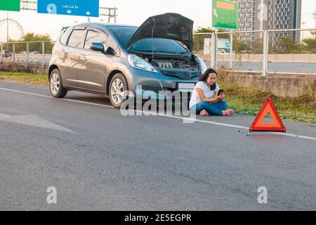 Asiatische Teenager-Frauen halten ein Handy zu Fuß rund um das Auto, stressige Stimmung während der Abendstunden. Entlang der Autobahn, weil ihr Auto brach tun Stockfoto