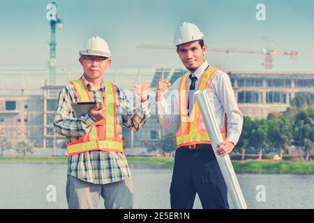 Zwei Techniker stehen auf der Baustelle Konstruktion glücklicher Erfolg, zwei Arbeiter halten Tablet und Blaupause Arbeitsplatz in der Nähe von Fluss Stockfoto
