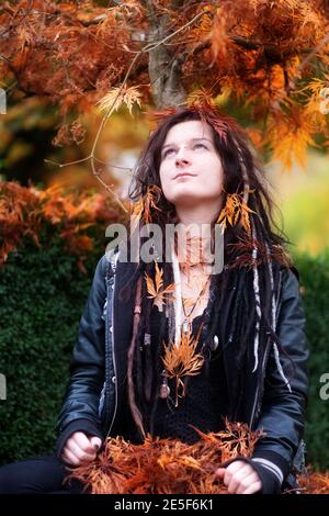 Schöne junge individuelle, exzentrische Frau, mit Dreadlocks, Piercing und Tattoo, mit schwarzer Lederjacke sitzt unter dem japanischen Ahornbaum und ist Stockfoto