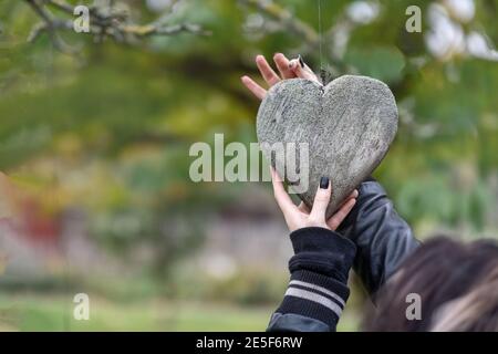 2 junge weibliche Hände mit schwarzen Fingernägeln befestigen ein mit Flechten bedecktes Holzherz auf einem Baum als Symbol für Liebe und Valentinstag, Raum kopieren Stockfoto