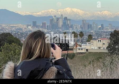 Carrie Giordano fotografiert den Vollmond, der über uns aufsteigt Die Skyline von Downtown Los Angeles mit dem schneebedeckten San Gabriel Berge als Kulisse Stockfoto