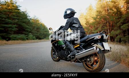 Motorradfahrer allein auf der Straße in einem Helm und Leder-Schutzausrüstung schaut zur Seite, sitzt auf einem schnellen Sport-Motorrad auf einem Wald-Backgro Stockfoto
