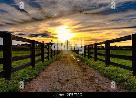 Schotterweg, der zu einer Scheune in der Ferne mit führt Holzschienenzäune auf beiden Seiten mit Sonnenuntergang Der Hintergrund Stockfoto