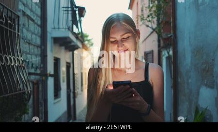 Nahaufnahme einer positiven blonden Frau, die etwas auf dem Smartphone liest, während sie auf der Straße läuft. Sie war glücklich und lächelte. Sommerzeit. Stockfoto