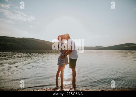 Volle Länge des Paares in der Liebe küssen am Strand bei Sonnenuntergang. Portrait von jungen Mann und Frau Umarmung und küssen am See an schönen Sommerabend Stockfoto