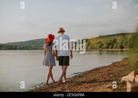 Rückansicht eines Paares, das die Hände hält und bei Sonnenuntergang am Strand entlang läuft. Rückansicht von Mann und Frau, die am Sommerabend am See spazieren gehen. Stockfoto