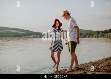 Vorderansicht eines glücklichen Paares, das die Hände hält und bei Sonnenuntergang am Strand entlang läuft. Voller lächelnder Mann und Frau, die auf der Summe am See spazieren gehen Stockfoto