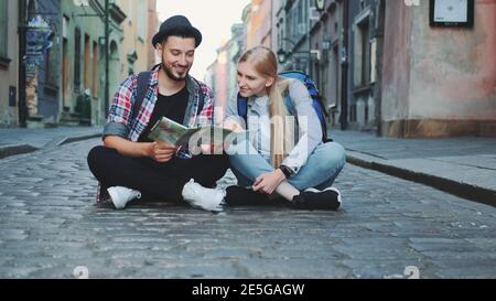 Junge Touristen pärchen mit Karte, sitzen auf dem Bürgersteig und bewundern die historische Umgebung. Sie sind aufgeregt und lächeln. Stockfoto