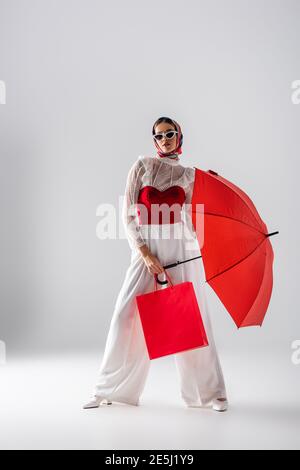 In voller Länge modische Frau in Kopftuch und Sonnenbrille halten Roter Regenschirm und Einkaufstasche beim Posieren auf Weiß Stockfoto