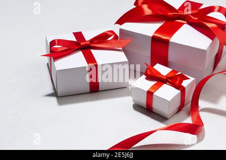 Weiße Geschenkschachteln in verschiedenen Größen mit rotem Seidenband auf weißem Holzbrett als Bordüre, Nahaufnahme, Hochzeitshintergrund. Stockfoto