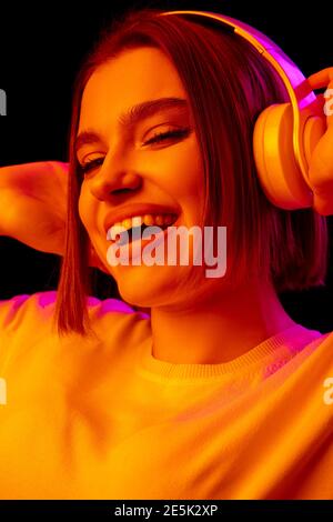 Glücklich, verrückt. Kaukasische Frau Porträt auf schwarzem Studio-Hintergrund in rosa-orange Neonlicht. Schönes weibliches Modell mit Kopfhörern. Konzept der menschlichen Emotionen, Gesichtsausdruck, Verkauf, Werbung, Mode. Stockfoto