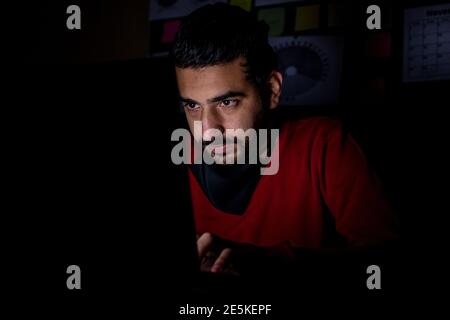 Junger Mann in der Überstunden im Dunkeln von seinem Office arbeitet hart an seinem Projekt mit einem ernsten Blick Auf seinem Gesicht Stockfoto