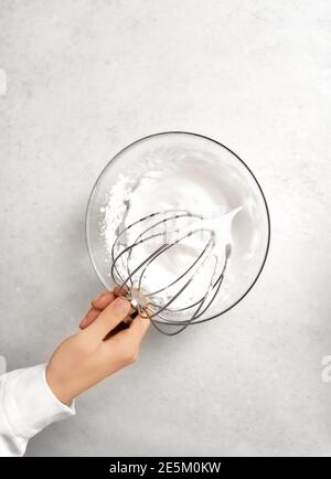 Die Hand der jungen Frau, die den Schneebesen über die Schüssel mit Schlagsahne hält. Weißer Küchenhintergrund. Essen und hausgemachtes kulinarisches Konzept. Flache Lay-Optik. Stockfoto