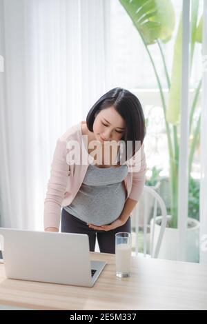 Junge asiatische schwangere Frau, die von zu Hause aus mit Computer und Trinken Milch für die gute Gesundheit ihres Babys Stockfoto