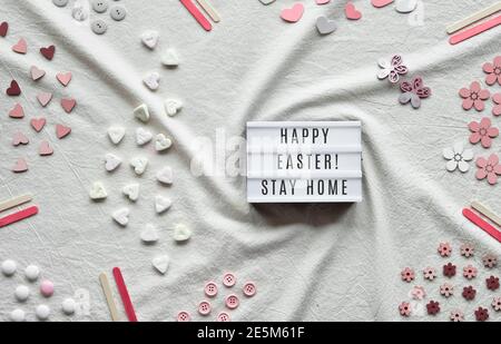 Happy Easter Stay Home Text auf Leuchtkasten. Flaches Lay mit Leuchtkasten auf Elfenbein, aus weißem Textil. Herzen, Süßigkeiten, Knöpfe, verschiedene Dekorationen auf angeordnet Stockfoto