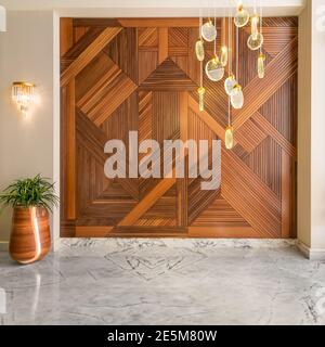 Halle mit Holzverkleidungen Wand mit geometrischen Mustern, hohen runden Holzpflanzer mit grünen Büschen, zeitgenössischen Glas LED hohen Kronleuchter und weißen Marmorboden in modernen Haus dekoriert Stockfoto