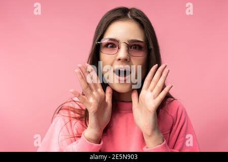 Foto von attraktiven jungen Frau in Brille, niedlich und schockiert Gesicht. Trägt lässige rosa T-Shirt weiße Hose isoliert rosa Farbe Hintergrund Stockfoto