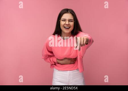 Foto von attraktiven jungen Frau in Brille lacht zeigt auf Kamera, trägt beiläufige rosa T-Shirt weiße Hose isoliert rosa Farbe Hintergrund Stockfoto