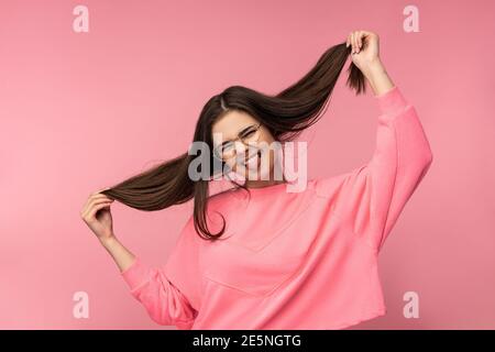 Foto von attraktiven Frau in der Brille Spaß hält Haare Tragen Sie casual rosa T-Shirt weiße Hose isoliert rosa Farbe Hintergrund Stockfoto