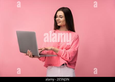 Foto von attraktiven Dame in der Brille mit Online-Meeting, hält Laptop und arbeitet online. Tragen beiläufige rosa T-Shirt weiße Hose isoliert rosa Farbe Stockfoto