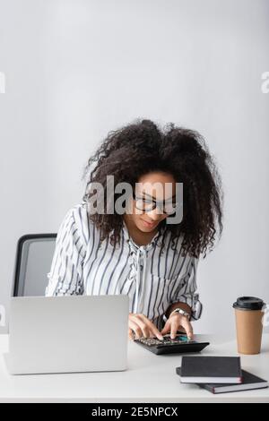 afroamerikanischer Freiberufler in Brillen mit Taschenrechner, während Zählen in der Nähe Laptop und Papiertasse auf dem Schreibtisch Stockfoto