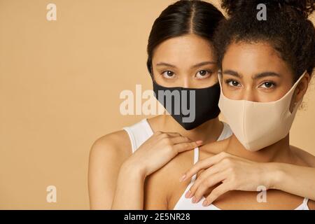 Zwei attraktive junge Frauen, weibliche Freunde tragen schützende Gesichtsmasken, während zusammen posieren isoliert auf beigem Hintergrund. Sicherheit, Pandemiekonzept Stockfoto