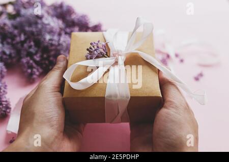 Hände halten Geschenkbox mit Band und Flieder Blumen auf rosa Papier. Happy Mothers Day und valentinstag Glückwünsche Konzept. Lila Fliederblüte Stockfoto