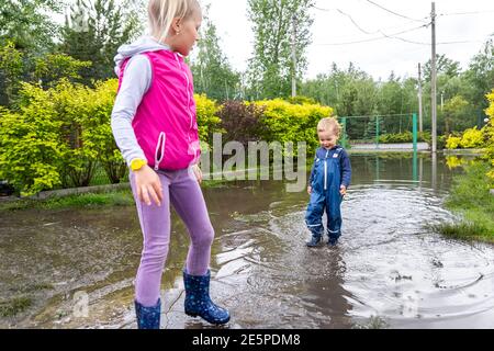 Paar von zwei niedlichen blonde kaukasischen kleinen Kindern Bruder und Schwester genießen haben Spaß spielen springen in schmutzige Pfütze tragen Blaue wasserdichte Hose und Stockfoto