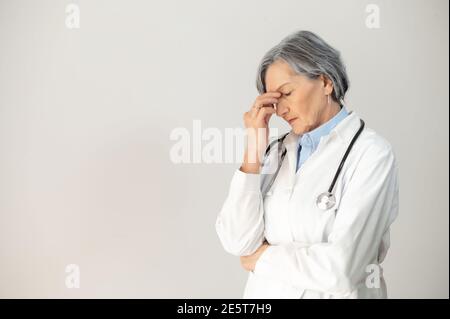 Gestresste ältere Ärztin mit einem Stethoskop in einem medizinischen Gewand, das ihr Gesicht bedeckt, sich überwältigt fühlt, erhielt schlechte verheerende Nachrichten, erfahren Stockfoto