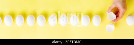 Weiße rohe Hühnereier liegen in Reihe auf einem gelben trendigen Hintergrund und weibliche Hand hält gebrochenes Ei. Ostern minimal kreative Komposition. Spring Happy Stockfoto